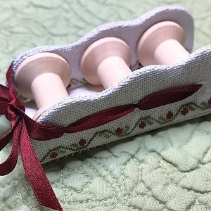 May include: A white fabric spool holder with three wooden spools. The holder is decorated with a red ribbon and a green and red embroidered design.