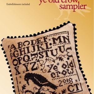 May include: A cross-stitch sampler featuring a black crow design with the text "ye old crow, sampler". The sampler includes the alphabet and the year 2018. The sampler is framed with a black pom-pom trim.