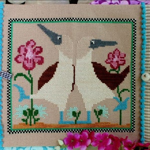 May include: A handmade, cross-stitched book cover featuring two white birds facing each other, surrounded by pink flowers and blue accents. The cover is tan with a black and white checkered border and a blue pom-pom trim. The text "Lindy Stitches" is in the lower right corner.