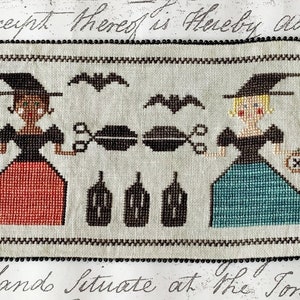 May include: Cross-stitch pattern featuring two witches with black hats and dresses, one with a red skirt and the other with a blue skirt. They are holding scissors and a cauldron. The background is white with black bats.