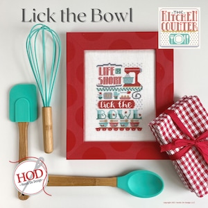 Könnte beinhalten: Ein rot gerahmter Kreuzstich mit dem Text "Life is Short Lick the Bowl" umgeben von Küchenmotiven. Der Kreuzstich ist auf weißem Hintergrund mit roten Punkten. Im Bild sind Küchenutensilien, ein rot-weiß kariertes Geschirrtuch und ein Logo für "The Kitchen Counter" zu sehen.