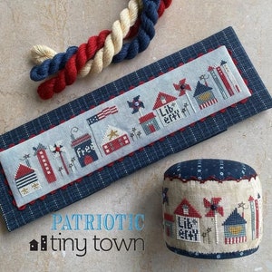 May include: A cross-stitch sampler with a patriotic theme. The sampler features a row of houses with red, white, and blue accents. The text "PATRIOTIC tiny town" is printed below the sampler.