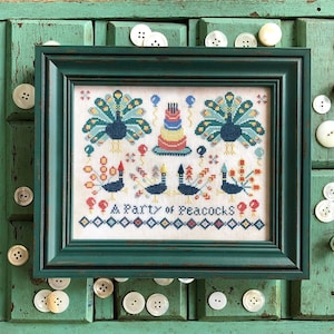 May include: A framed cross-stitch design featuring a party of peacocks with a birthday cake and balloons. The design is in a white frame with a teal green border. The text "A Party of Peacocks" is stitched below the design.