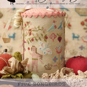 May include: A cream-colored pincushion with a pink ruffled top and a cross-stitch design of a tree with birds and flowers. The text "Five Songbirds Pin Drum" is below the pincushion.