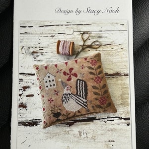 May include: Cross-stitch pattern for a pincushion featuring a rooster with a red comb and a white and black striped tail. The pattern is titled "Le Poulet Pinkeep" and is designed by Stacy Nash.