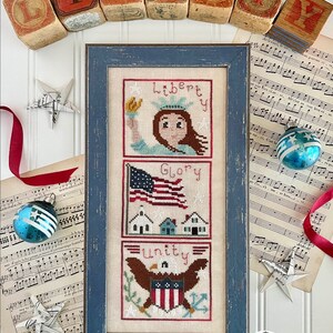 May include: A framed cross-stitch sampler with three panels depicting a woman holding a torch, an American flag, and an eagle. The panels are labelled "Liberty", "Glory", and "Unity". The sampler is surrounded by wooden blocks spelling "LIBERTY" and other patriotic decorations.