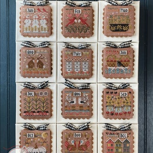 May include: Twelve framed cross-stitch designs, each depicting a different month of the year. The frames are brown with a scalloped edge and a black and white checkered ribbon. The designs are colorful and feature various seasonal themes, such as snowmen, birds, flowers, and pumpkins. The text "Season 2 A Year of Celebrations continues" is at the top of the image.