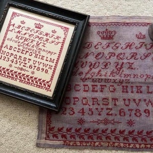 Op de afbeelding: Een ingelijste kruissteek sampler met een zwarte lijst, met rode letters en een bloemenrand. De sampler toont het alfabet, cijfers en de naam "MARTHA MASIALERS/89A". Daarnaast is een vergelijkbare, niet-ingelijste sampler zichtbaar.