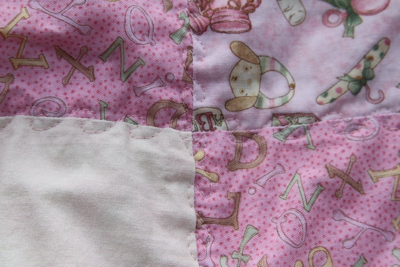 Puede incluir: Una colcha de retazos rosa y blanca con un borde festoneado. La colcha est&aacute; hecha de diferentes telas con varios patrones, incluyendo letras y peque&ntilde;as ilustraciones.