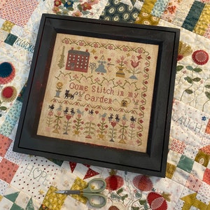 Può includere: Opera d'arte a punto croce incorniciata con una cornice nera. L'opera presenta una scena di giardino con fiori, una casa e il testo "Come Stitch in my Garden". Un paio di forbici poggia sotto la cornice su una trapunta floreale.