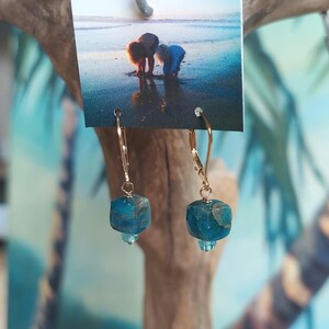 May include: Gold-colored drop earrings with blue-green, cube-shaped stones. The earrings are displayed against a backdrop of a beach scene and a wooden branch. The earrings have a small, round, blue-green stone dangling below the cube.