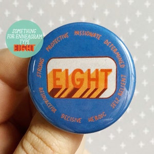 May include: Blue and orange button with the word "EIGHT" in large letters. The button has a white border and the words "Strong, Protective, Passionate, Determined, Resourceful, Decisive, Heroic, Self-Reliant" around the edge.