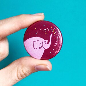 May include: A maroon button with a pink elephant design. The elephant is smiling and has confetti around it.