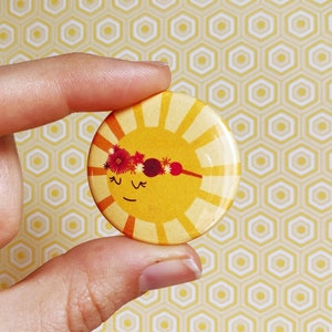 May include: A yellow sun with a smiling face and a red, white, and pink flower crown. The sun is on a white button with a yellow background.
