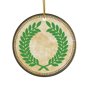 May include: Round ceramic ornament with a green laurel wreath design. The wreath is centered on a beige background with a decorative border. A gold string is attached for hanging.