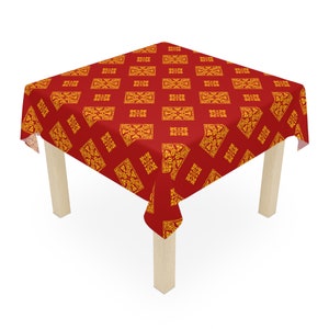 May include: A red tablecloth with a repeating gold pattern of ornate rectangular and square designs. The tablecloth is draped over a square table with light-colored wooden legs, creating a decorative and functional piece.