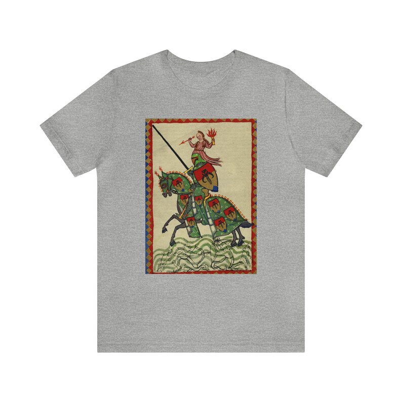 May include: A gray t-shirt featuring a medieval illustration of a knight on horseback. The artwork depicts a knight in armor with a lance, riding a horse with green and red accents. The design is framed with a red and blue border.