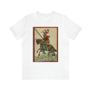 May include: White t-shirt featuring a medieval illustration of a knight on horseback. The artwork depicts a knight in red and green armor with a lance, riding a green horse with red and green accents. The background is beige with a red border.