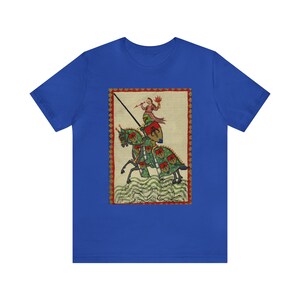 May include: Royal blue t-shirt featuring a medieval illustration of a knight on horseback. The artwork depicts a knight in armor with a lance, riding a horse with green and red accents. The background is a cream color with a red border.