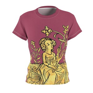 May include: A maroon t-shirt featuring a medieval-style illustration. The design depicts a woman holding a flower, rendered in yellow and black, set against a backdrop of stylized plants. The shirt has a crew neck and short sleeves.