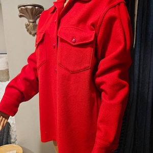 May include: A bright red wool shirt jacket with two chest pockets and button closures. The jacket has a classic collar and long sleeves. The garment is displayed on a mannequin.