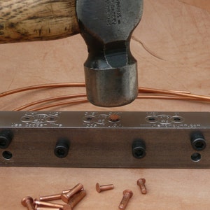 May include: A metal tool with three sets of holes for setting rivets. A hammer is being used to set a copper rivet. There are several copper rivets scattered around the tool.