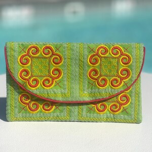 Può includere: Una pochette in tessuto verde con un design ricamato rosso e giallo. La pochette ha una chiusura a lembo e un bordo rosso.