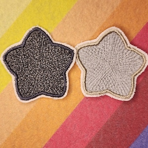 Könnte beinhalten: Zwei sternförmige, gestickte Aufnäher. Einer ist dunkelgrau mit weißem Rand und der andere hellgrau mit beigem Rand. Die Aufnäher befinden sich auf einem farbenfrohen, gestreiften Hintergrund aus Gelb, Orange, Rosa und Lila.