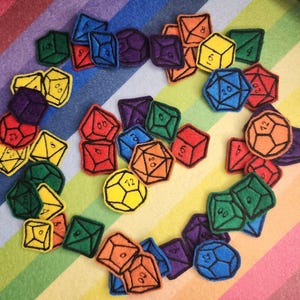 Può includere: Una collezione di dadi in feltro colorati di varie forme, tra cui cubi, d4, d8, d10, d12 e d20. Ogni dado è bordato di nero e presenta un colore diverso, come rosso, blu, verde, giallo, arancione e viola. I dadi sono disposti su uno sfondo a righe arcobaleno.