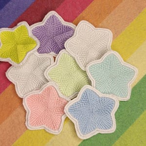May include: A selection of star-shaped coasters in various colours, including yellow, purple, white, green, pink, and blue. Each coaster has a textured surface and a white border, set against a rainbow striped backdrop.