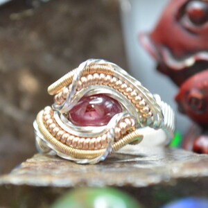 Puede incluir: Un anillo artesanal con una piedra preciosa roja central, intrincadamente envuelta en alambre plateado y dorado. El anillo se encuentra sobre una pequeña roca, rodeado de ópalos coloridos y un fondo borroso con una escultura de pez rojo y un gran cristal.