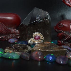 May include: A silver ring with a red gemstone set in a twisted band, surrounded by opals and other stones. The ring is displayed on a rock with a crystal behind it.