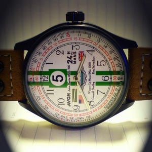 K&ouml;nnte beinhalten: Eine schwarz-wei&szlig;e Armbanduhr mit einem gr&uuml;n-wei&szlig;-roten Zifferblatt. Das Zifferblatt tr&auml;gt den Text "Avon Aston Martin DBRI/300 24h Mans" und "Winner Le Mans 24hr 1959 Carroll Shelby Roy Salvadori".
