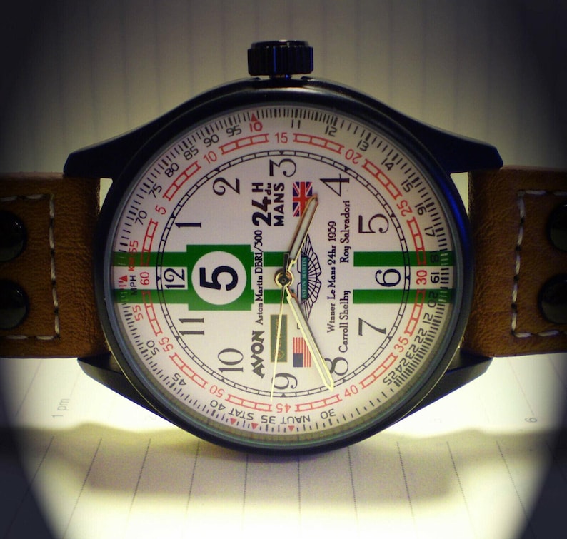 K&ouml;nnte beinhalten: Eine schwarz-wei&szlig;e Armbanduhr mit einem gr&uuml;n-wei&szlig;-roten Zifferblatt. Das Zifferblatt tr&auml;gt den Text "Avon Aston Martin DBRI/300 24h Mans" und "Winner Le Mans 24hr 1959 Carroll Shelby Roy Salvadori".