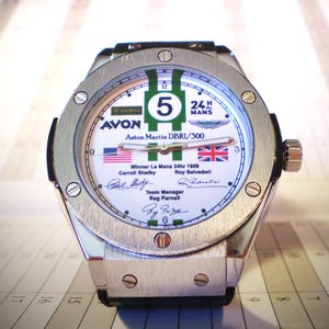 May include: A silver-toned wristwatch with a white face featuring green and white stripes, the number 5, and the text "24 MANS". The watch face includes the text "AVON" and "Aston Martin DBR1/300", along with the American and British flags.