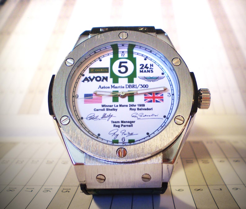 May include: A silver-toned wristwatch with a white face featuring green and white stripes, the number 5, and the text "24 MANS". The watch face includes the text "AVON" and "Aston Martin DBR1/300", along with the American and British flags.