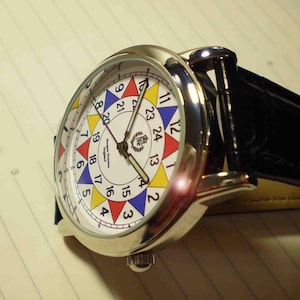 May include: A silver wristwatch with a white face and a black leather band. The face has a colorful design with numbers 1 through 24 around the edge. The watch has a small crown on the side.