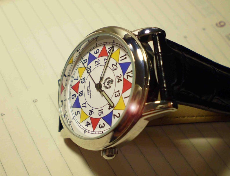 May include: A silver wristwatch with a white face and a black leather band. The face has a colorful design with numbers 1 through 24 around the edge. The watch has a small crown on the side.