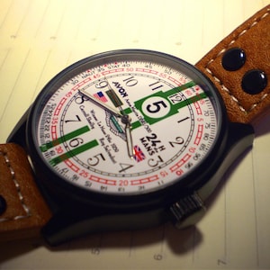 K&ouml;nnte beinhalten: Eine schwarz-wei&szlig;e Armbanduhr mit einem braunen Lederarmband. Das Zifferblatt der Uhr hat einen wei&szlig;en Hintergrund mit gr&uuml;nen, roten und blauen Akzenten. Das Zifferblatt der Uhr enth&auml;lt auch den Text "24H MANS" und "AVON D300".