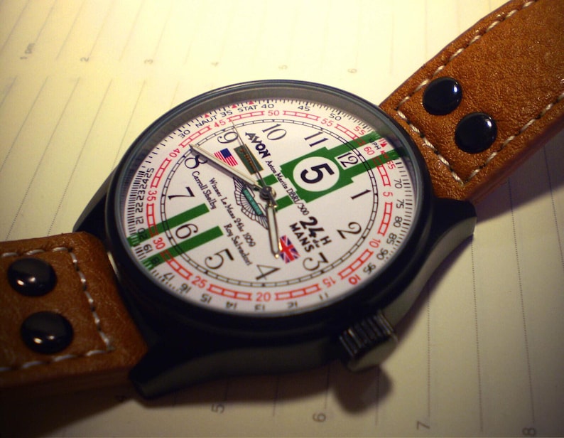 K&ouml;nnte beinhalten: Eine schwarz-wei&szlig;e Armbanduhr mit einem braunen Lederarmband. Das Zifferblatt der Uhr hat einen wei&szlig;en Hintergrund mit gr&uuml;nen, roten und blauen Akzenten. Das Zifferblatt der Uhr enth&auml;lt auch den Text "24H MANS" und "AVON D300".