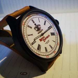 Puede incluir: Un reloj de pulsera con caja negra mate y correa de cuero color canela. La esfera blanca presenta números romanos negros, un logotipo rojo de Royal Enfield y un pequeño escudo en la parte superior. El borde exterior muestra marcas de 25 a 300.