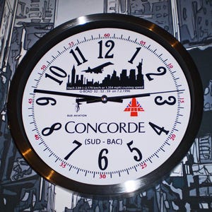 May include: A white clock with black numbers and hands, featuring a black and white image of the Concorde supersonic jet with the text "CONCORDE (SUD - BAC)" and "Mach 2.04 (-2,179 km/h or 1,354 mph) cruising speed G-BOAD 02:52:59 on 7.2.1996" printed on the face.