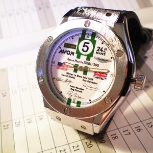 May include: A silver-toned wristwatch with a white face featuring green stripes and the number 5. The watch face includes text and flags, with a black leather band. The watch is on a white background.