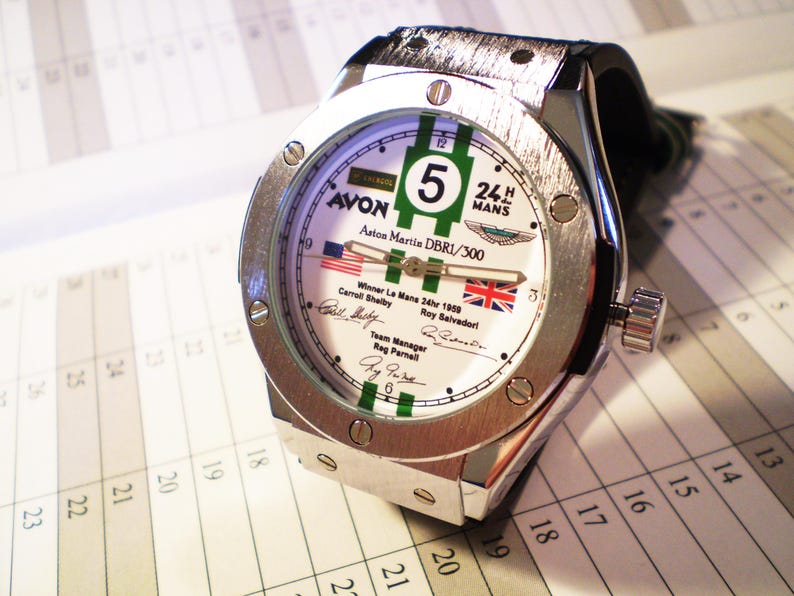 May include: A silver-toned wristwatch with a white face featuring green stripes and the number 5. The watch face includes text and flags, with a black leather band. The watch is on a white background.