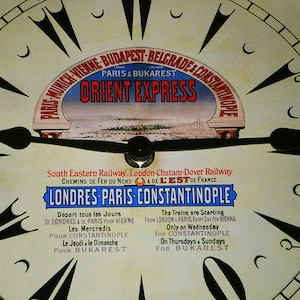 Puede incluir: Una esfera de reloj vintage con agujas negras y números romanos. La esfera del reloj es blanca con una pancarta azul y roja que dice "Orient Express" y "Londres París Constantinopla". La pancarta también incluye el texto "South Eastern Railway. London-Chatam-Dover Railway. Chemins de Fer du Nord & de L'Est de France."