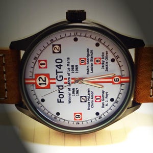 May include: A white watch face with black hands and black markings. The watch face has the text "Ford GT40" and "Winner of 24hrs of Le Mans" with the years 1966, 1967, 1968, and 1969. The watch face also has the names of the drivers who won the race in those years.