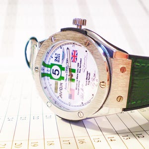 May include: A silver wristwatch with a white face featuring green and white racing stripes, the number 5, and the text "24 H MANS". The watch has a green leather band and is set against a calendar.
