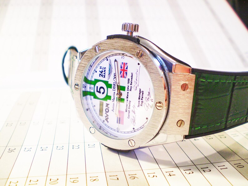 May include: A silver wristwatch with a white face featuring green and white racing stripes, the number 5, and the text "24 H MANS". The watch has a green leather band and is set against a calendar.