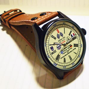 Könnte beinhalten: Eine schwarze Armbanduhr mit einem braunen Lederarmband. Das cremefarbene Zifferblatt zeigt schwarze römische Ziffern und den Text "GRAND PRIX DE L.A.C.F. REIMS". Die Uhr hat auch eine Zielflagge und eine französische Flagge.
