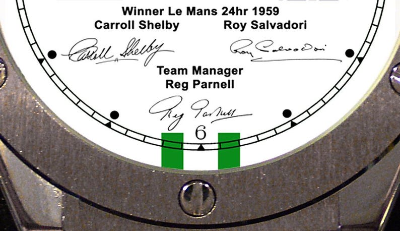 May include: Close-up of a watch face with the text "Winner Le Mans 24hr 1959" and the names Carroll Shelby, Roy Salvadori, and Reg Parnell. The watch face is white with black markings and green accents, surrounded by a silver metal bezel.
